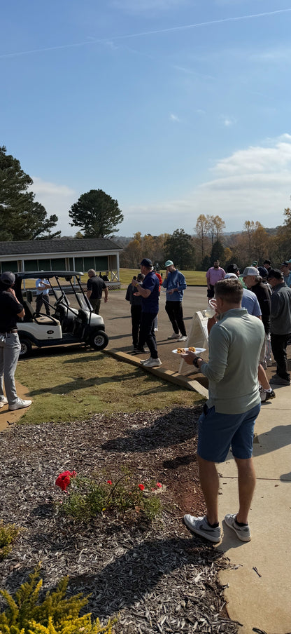 Group of people standing outdoors with golf carts and a clear sky at On-Brand Open charity golf tournament