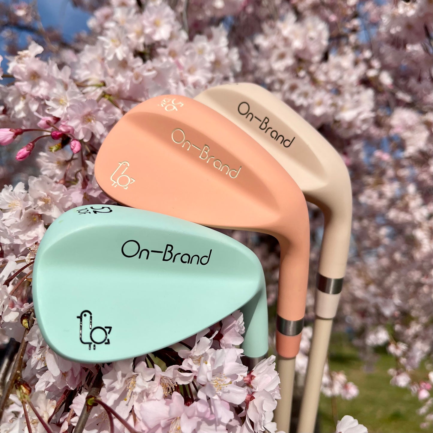 Two golf clubs, one peach and one green, with 'On-Brand' text against a cherry blossom background.