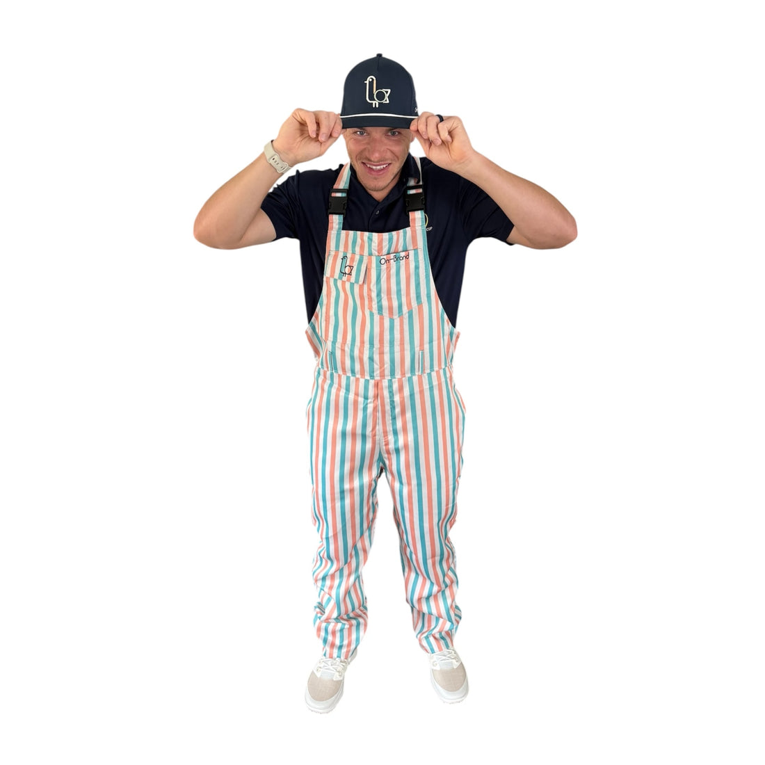 Golfer in On-Brand Golf Pair-a-Keets peach, seafoam, and cream striped bib overalls tipping a navy seagull cap, front view
