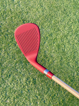 Burgundy Phoenix Wedge at address on lush fairway turf, grooves visible – On-Brand Golf