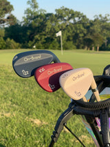 Three Phoenix Wedges — cream 50°, burgundy 54°, navy 58° — in a golf bag on the course with a pin flag behind